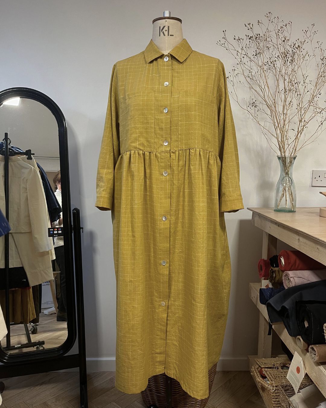 Yellow checkered dress on a mannequin against a white wall with a mirror and shelves in the background.