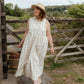 Woman in a white dress and hat standing near a wooden gate with a basket, outdoors.