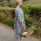 Woman in a blue checkered dress holding a basket with flowers on a rural road.