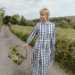 Woman in a blue checkered dress holding a basket of flowers on a rural road.
