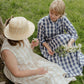Two women sitting on grass, one in a natural check  dress and straw hat, the other in a blue checkered dress, holding flowers.
