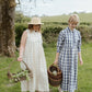 Two women in a garden holding picnic baskets, one in a natural check dress and straw hat, the other in a blue checkered dress.