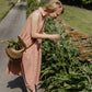 Woman in a pink dress tending to plants by a road with a house in the background
