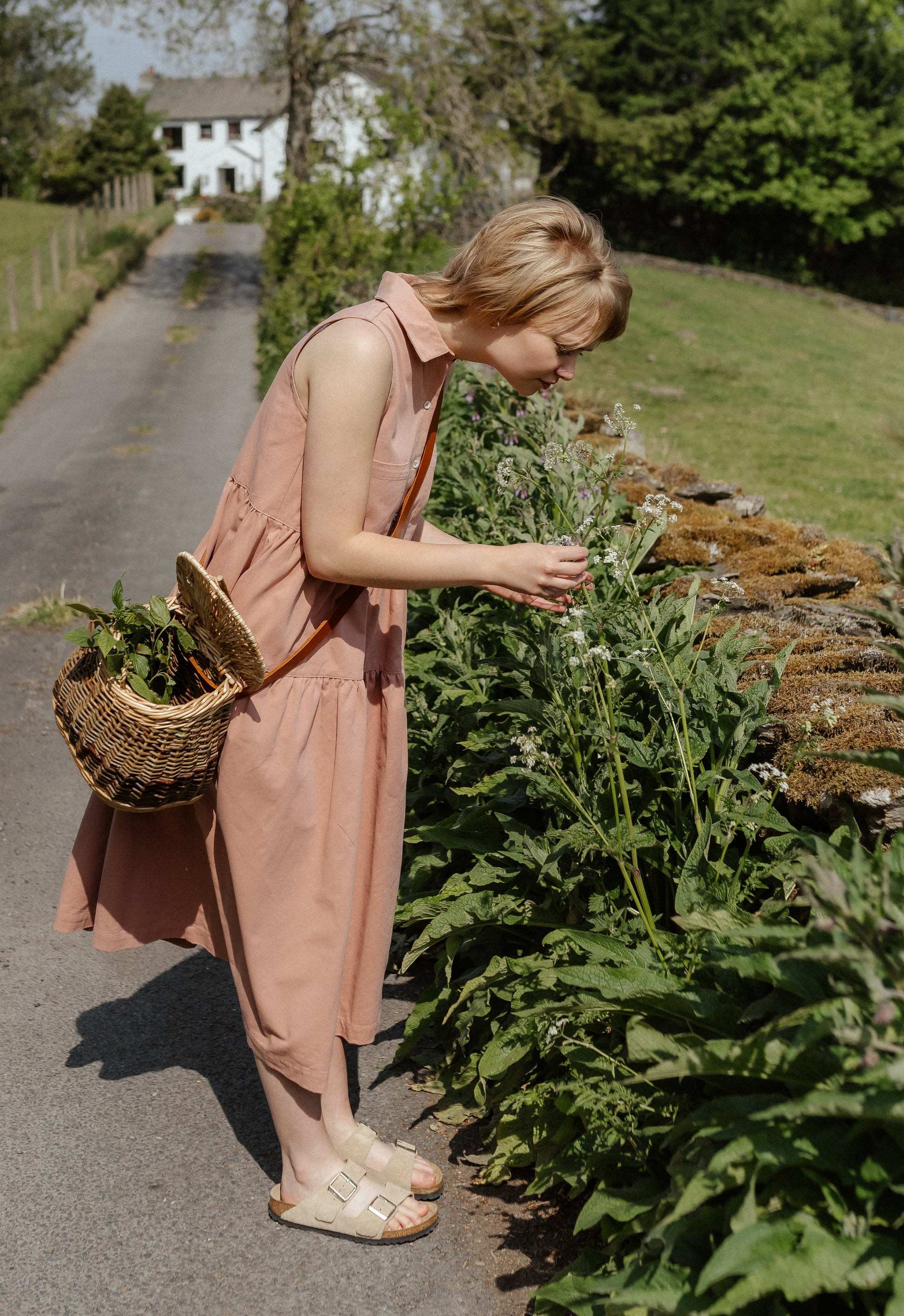 Woman in a pink dress tending to plants by a road with a house in the background