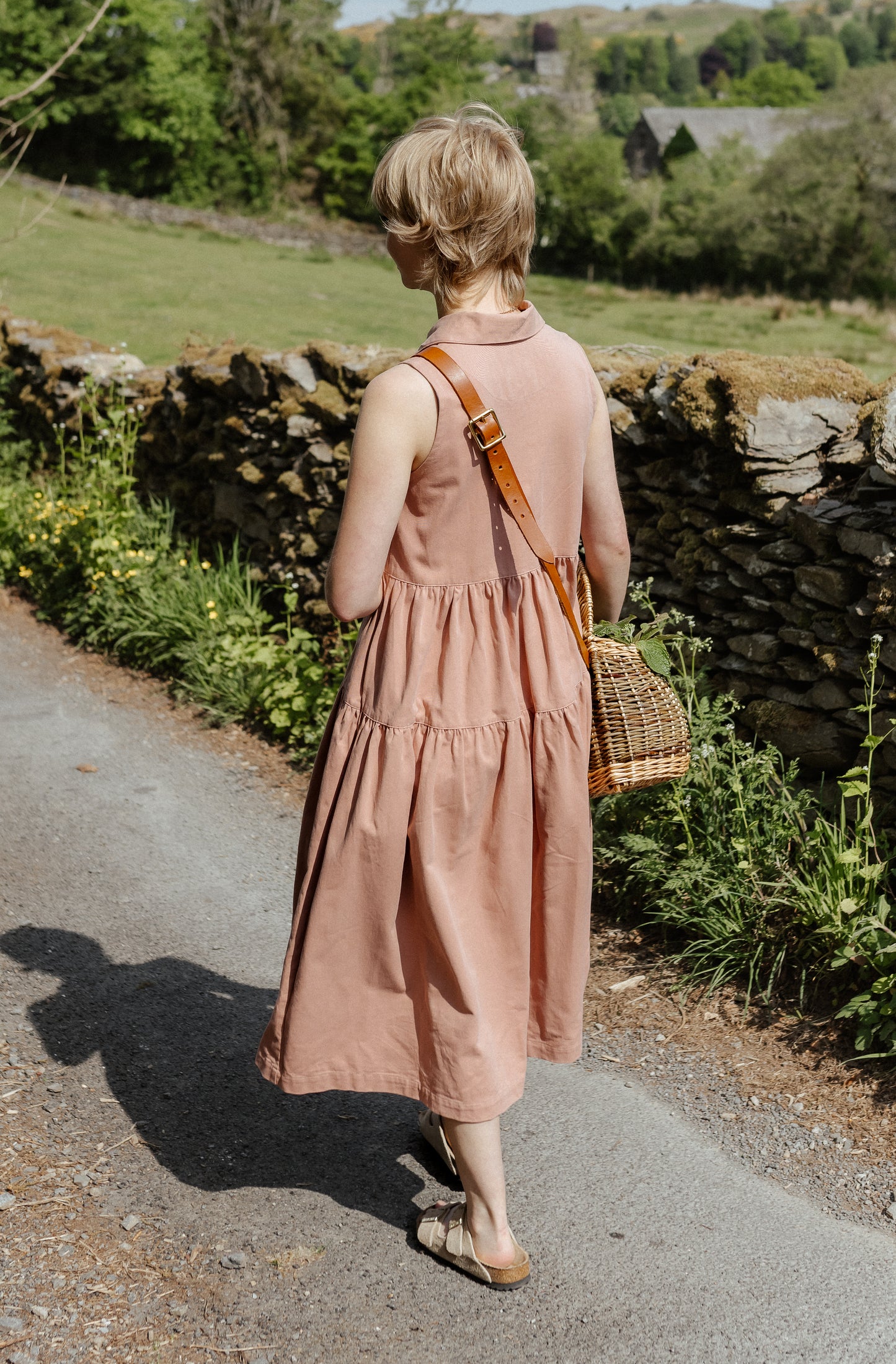 woman standing on road in country setting wearing a sleeveless pink dress.