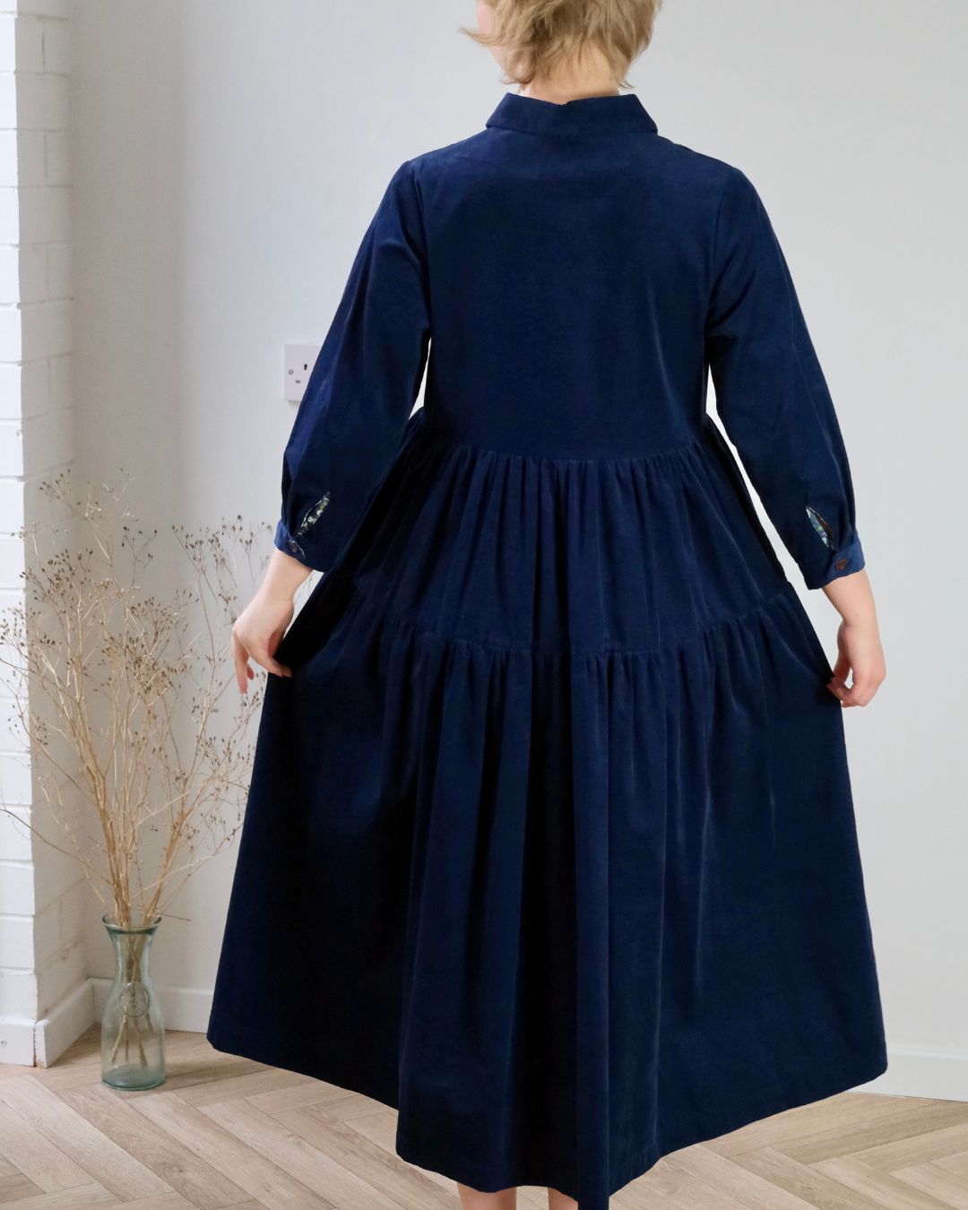 Person wearing a navy blue dress standing in a room with a white wall and wooden floor.