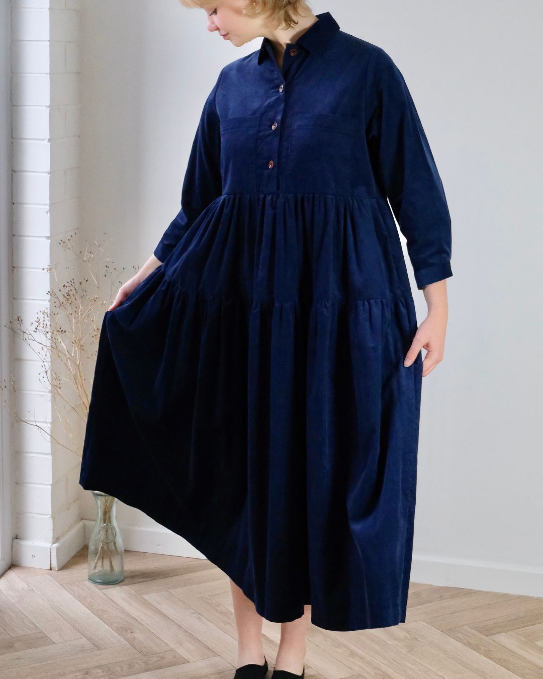 Person wearing a navy blue dress standing indoors with a light gray wall and wooden floor.