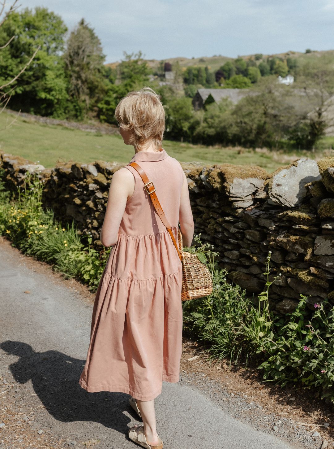 Woman in Effie pink dress walking along a country path with stone walls and greenery.