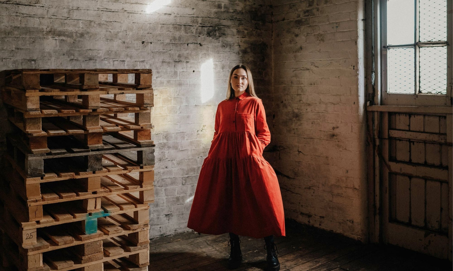 Woman in a red dress standing in an industrial setting with wooden pallets and brick walls.