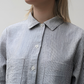 A woman standing in a rustic location wearing a gray peplum shirt with long sleeves and a collar, the shirt has a button-through design and frill detailing.