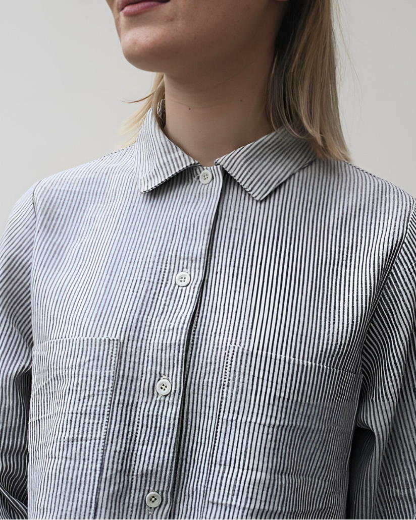 A woman standing in a rustic location wearing a gray peplum shirt with long sleeves and a collar, the shirt has a button-through design and frill detailing.