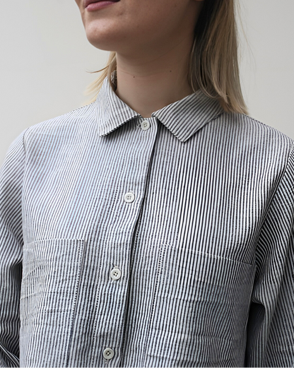 A woman standing in a rustic location wearing a gray peplum shirt with long sleeves and a collar, the shirt has a button-through design and frill detailing.