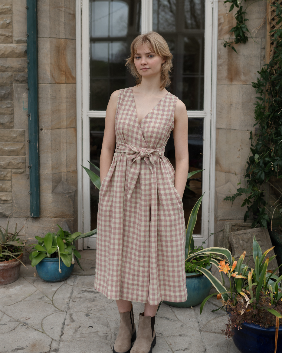 A woman standing outdoors wearing a beige and pink checkered sleeveless wrap dress with a tie at the waist.