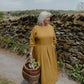 Woman in a yellow dress holding a basket with plants, standing in front of a stone wall.