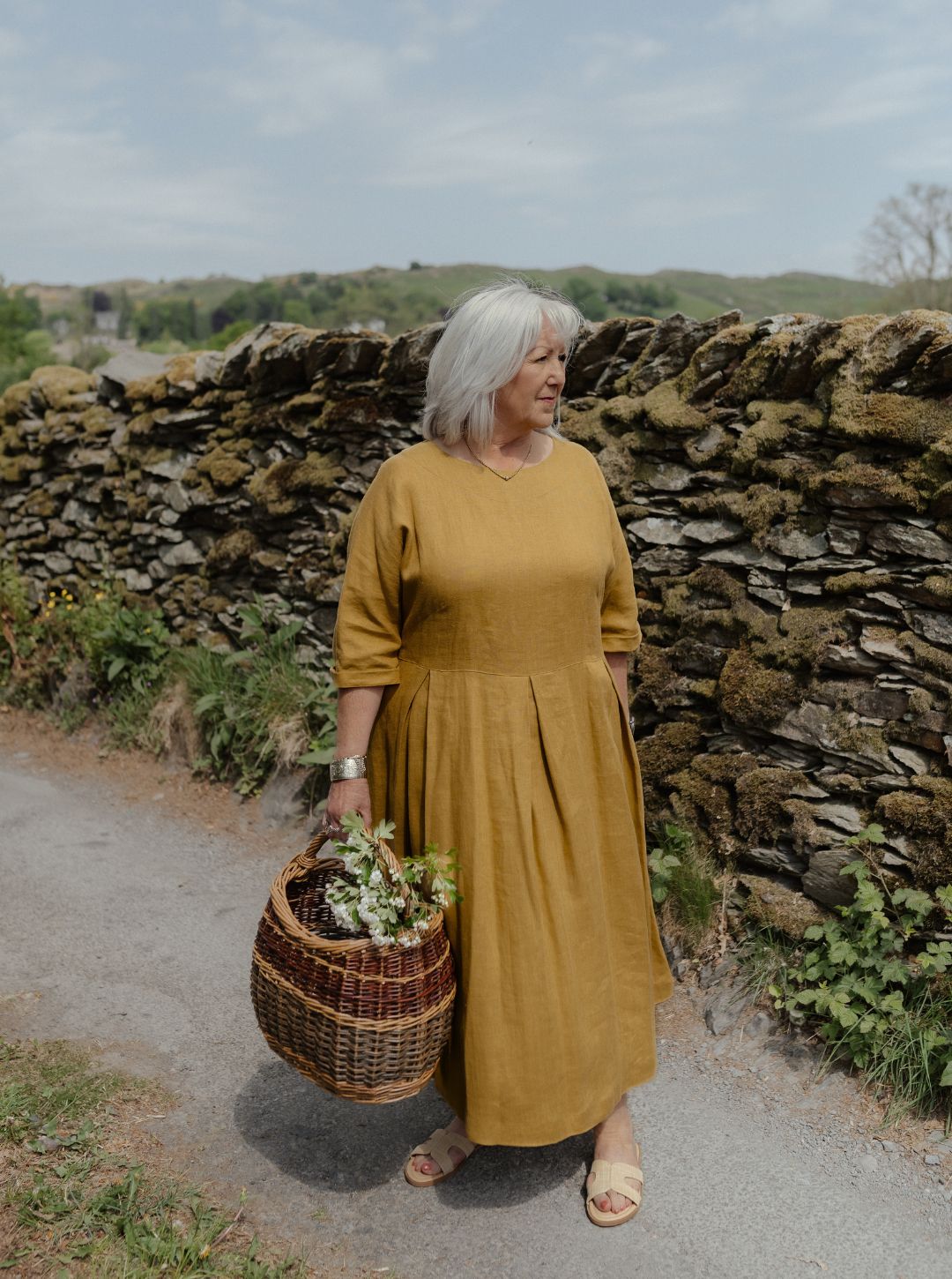 Woman in a yellow dress holding a basket with plants, standing in front of a stone wall.