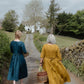 Two women walking away on a path in a rural setting, one in a blue dress and the other in a yellow dress.