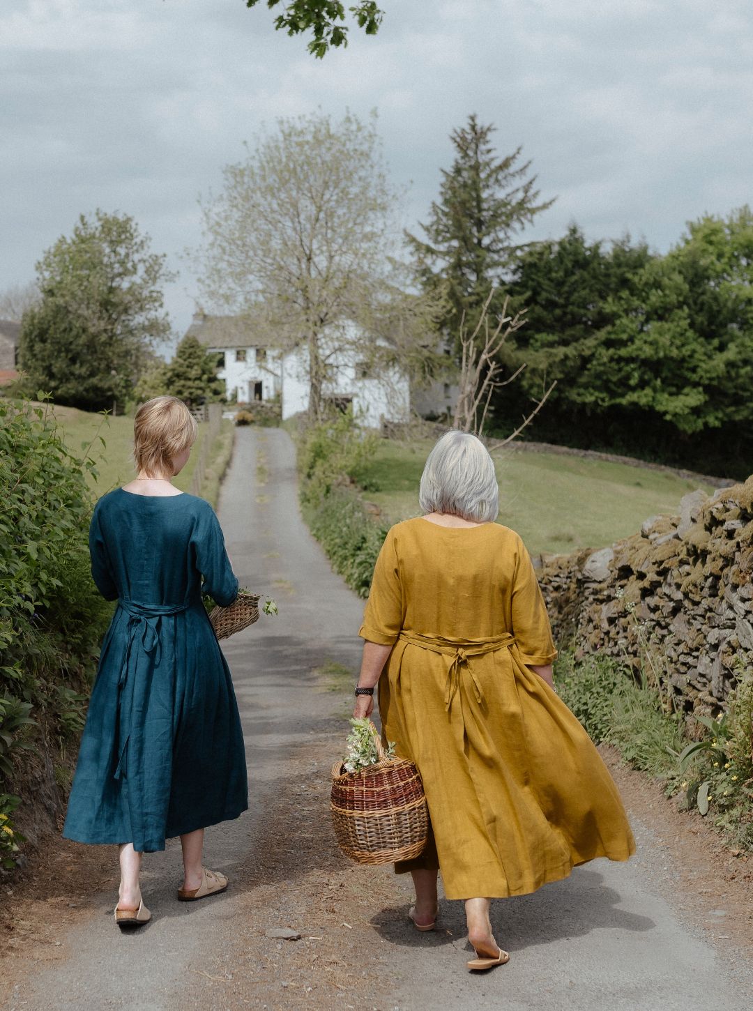 Two women walking away on a path in a rural setting, one in a blue dress and the other in a yellow dress.