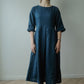 A woman wearing a dark teal blue linen midi dress with three-quarter sleeves, standing in front of a plain light gray wall on a light wood herringbone floor.