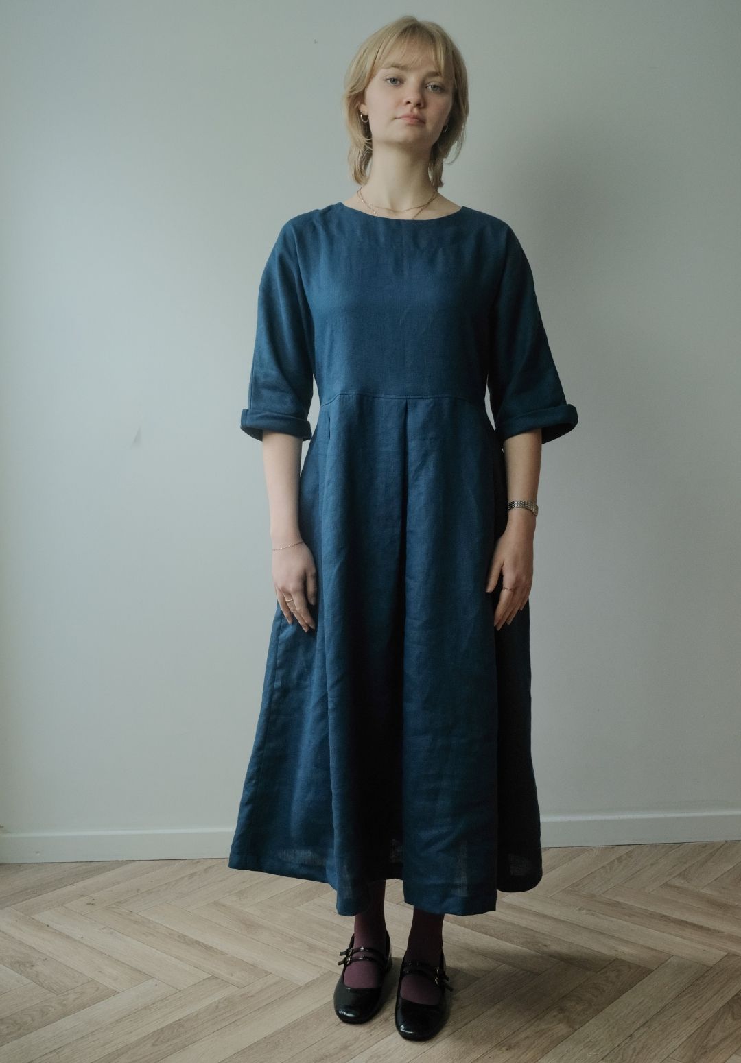 A woman wearing a dark teal blue linen midi dress with three-quarter sleeves, standing in front of a plain light gray wall on a light wood herringbone floor.