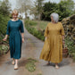 Two women walking outdoors, one in a teal dress and the other in a mustard yellow dress, with a rustic background.