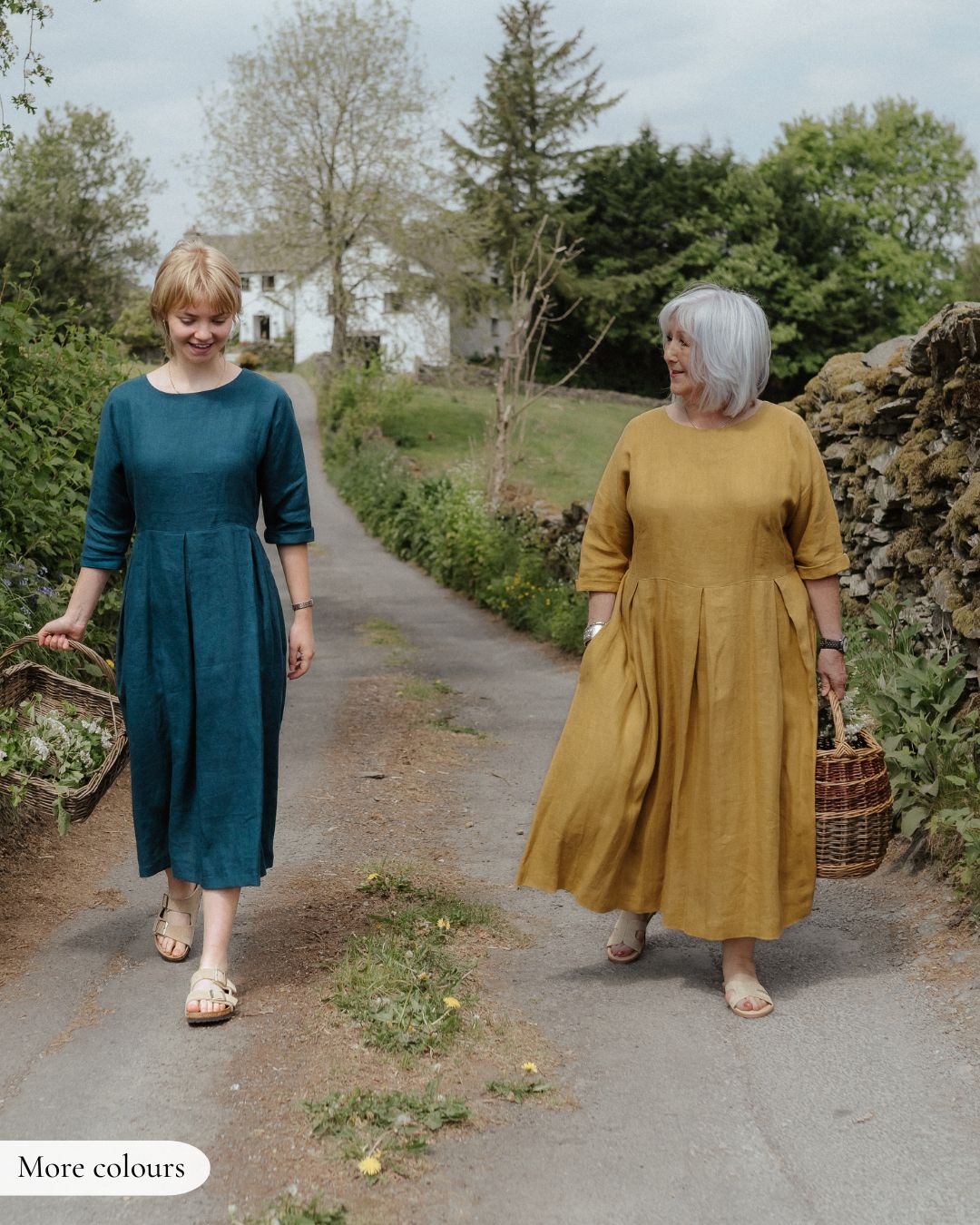 Two women walking outdoors, one in a teal dress and the other in a mustard yellow dress, with a rustic background.