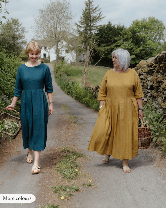 Two women walking outdoors, one in a teal dress and the other in a mustard yellow dress, with a rustic background.
