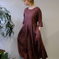 A woman wearing a loose, dusty rose-brown linen midi dress with three-quarter sleeves and a pleated waist, standing in a room with a light gray wall and wooden floor next to a green plant.