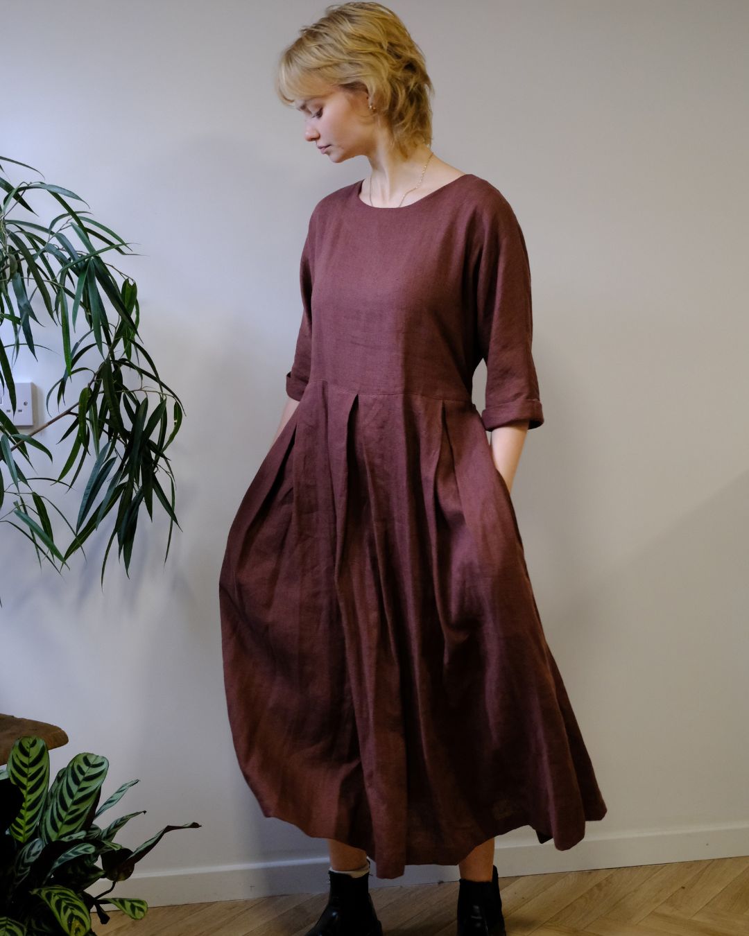 A woman wearing a loose, dusty rose-brown linen midi dress with three-quarter sleeves and a pleated waist, standing in a room with a light gray wall and wooden floor next to a green plant.