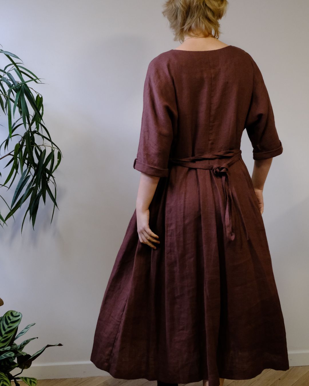 A woman viewed from behind wearing a dark brown linen dress with three-quarter sleeves and a tie belt at the back, standing in an indoor setting with a light gray wall and a green plant on the left.