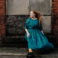 A woman wearing a teal green midi dress with three-quarter sleeves and a tie waist sits on a stone ledge in front of a brick wall and metal window.
