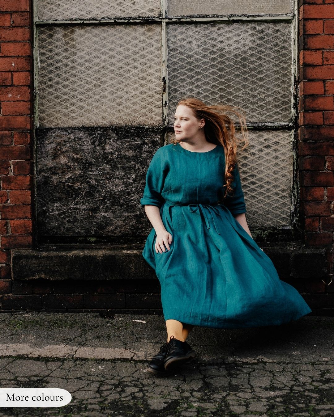 A woman wearing a teal green midi dress with three-quarter sleeves and a tie waist sits on a stone ledge in front of a brick wall and metal window.