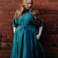 A model wearing a blue  linen dress with elbow length  sleeves, standing against a brickwall