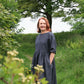 Woman in a long dark dress standing in a natural setting with trees and greenery.