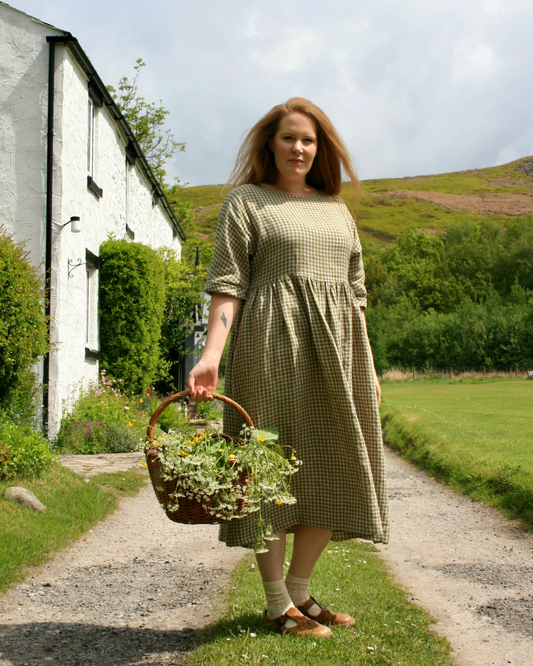 A woman standing in an outdoor setting wearing a green  linen dress with a scooped neckline, rolled sleeves, and a full skirt.