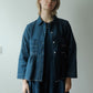 A woman with short blonde hair wearing a dark indigo denim jacket over a matching dark blue dress, standing against a plain light gray wall.§