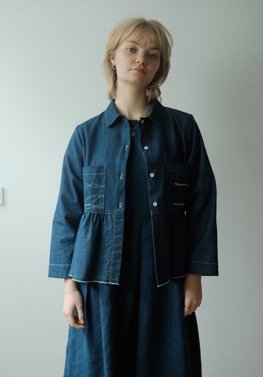 A woman with short blonde hair wearing a dark indigo denim jacket over a matching dark blue dress, standing against a plain light gray wall.§