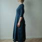 A woman stands in profile wearing a dark blue linen midi dress with three-quarter sleeves and a tie at the waist, paired with dark tights and black loafers, set against a plain light gray wall and wooden parquet floor.