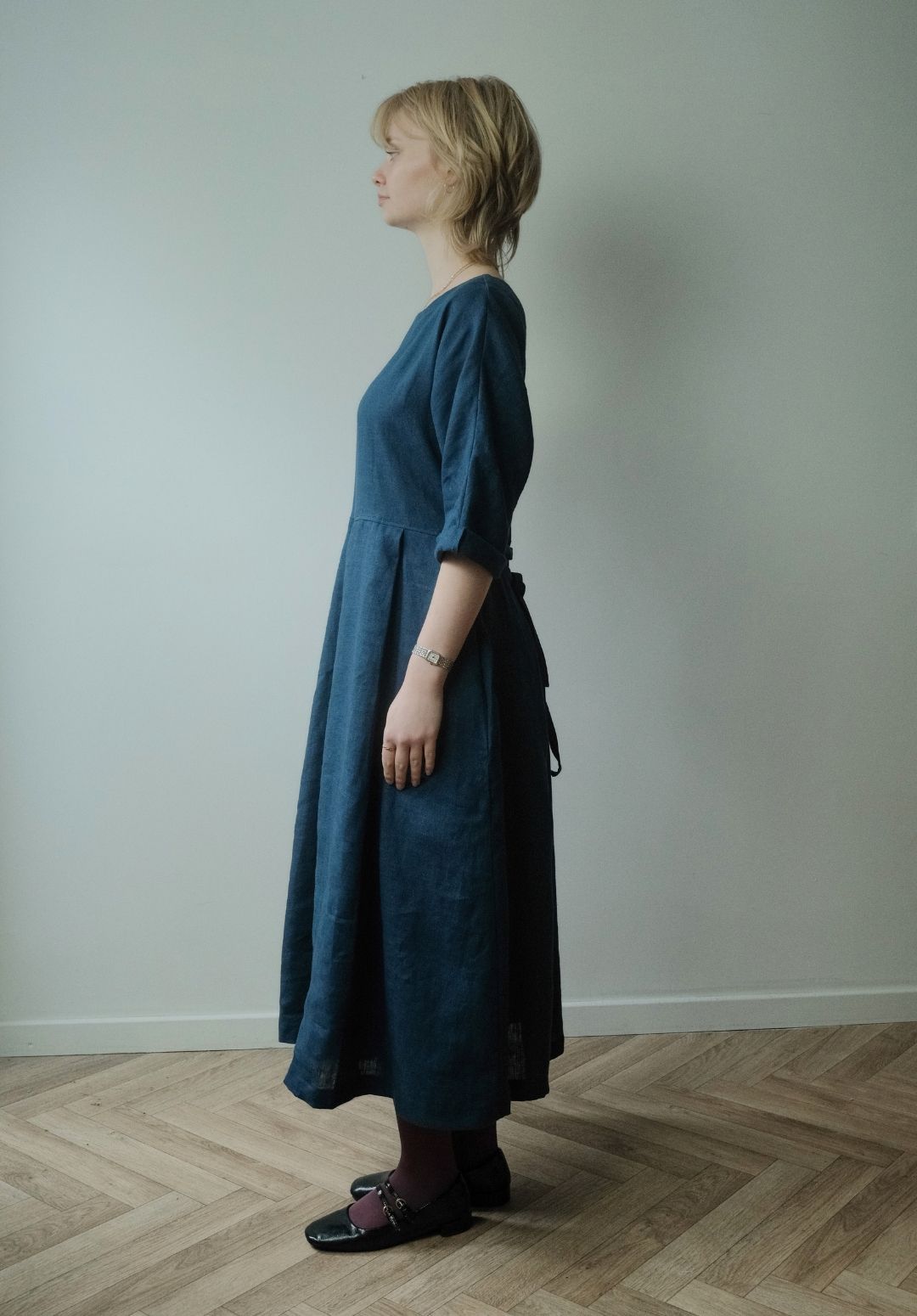 A woman stands in profile wearing a dark blue linen midi dress with three-quarter sleeves and a tie at the waist, paired with dark tights and black loafers, set against a plain light gray wall and wooden parquet floor.