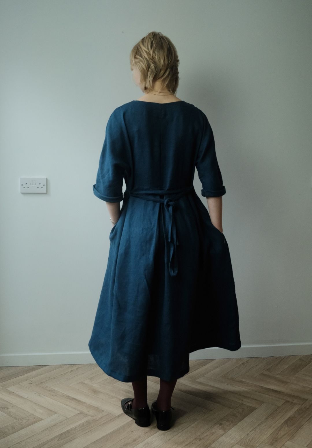 A woman viewed from behind wearing a dark teal blue linen midi dress with three-quarter sleeves and a tie waist, standing on a light wood herringbone floor against a plain light gray wall.