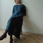 A woman with blonde hair sits on a wooden chair in a minimalist room with a light-colored wall and herringbone wood floor, wearing a dark blue linen midi dress, dark purple tights, and black leather shoes.
