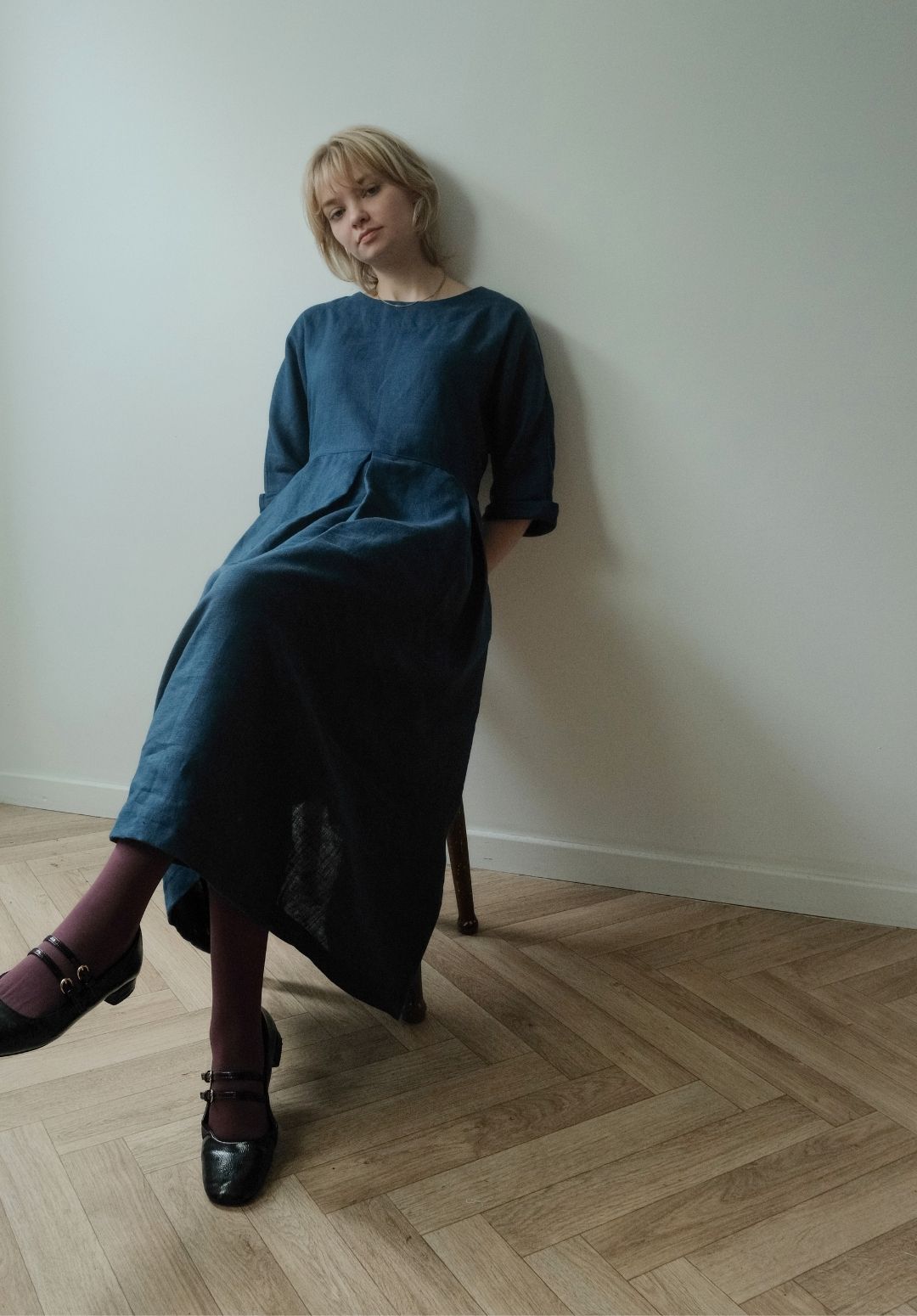 A woman with blonde hair sits on a wooden chair in a minimalist room with a light-colored wall and herringbone wood floor, wearing a dark blue linen midi dress, dark purple tights, and black leather shoes.