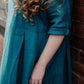A woman wearing a teal blue linen dress, leaning against a red brick wall. The dress has three-quarter sleeves with rolled cuffs and a gathered waist seam.
