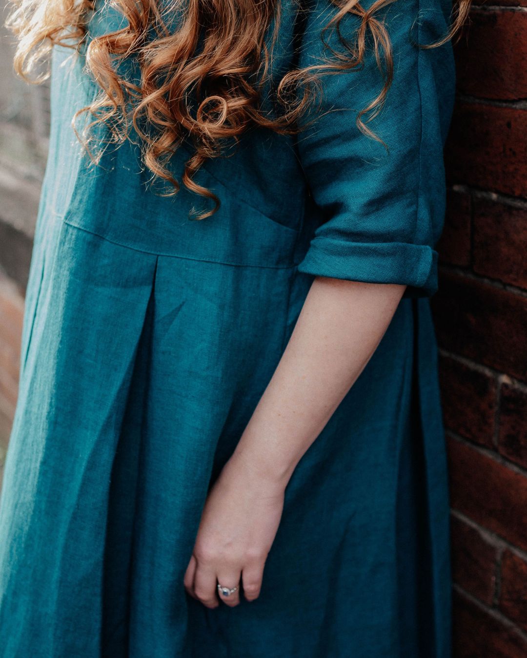A woman wearing a teal blue linen dress, leaning against a red brick wall. The dress has three-quarter sleeves with rolled cuffs and a gathered waist seam.