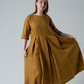 Woman standing against white background wearing a gold coloured linen dress with pleats.