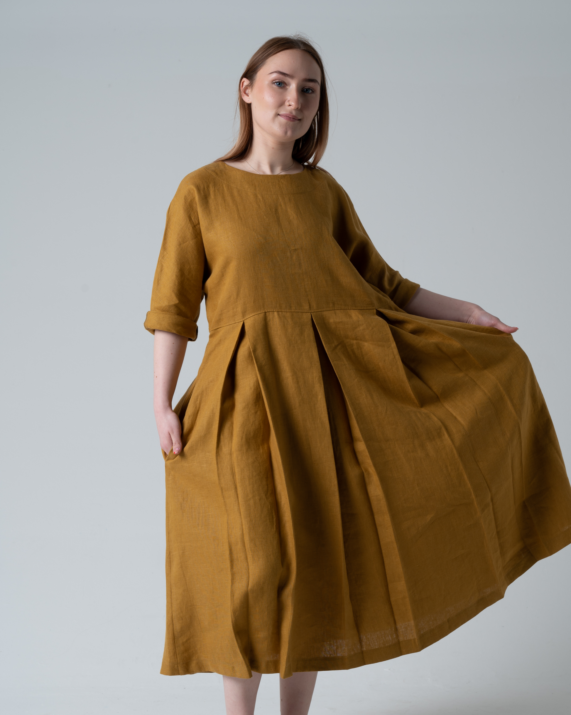 Woman standing against white background wearing a gold coloured linen dress with pleats.