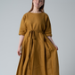 A model wearing golden linen dress with elbow length sleeves and pleated skirt. Standing against a white background.