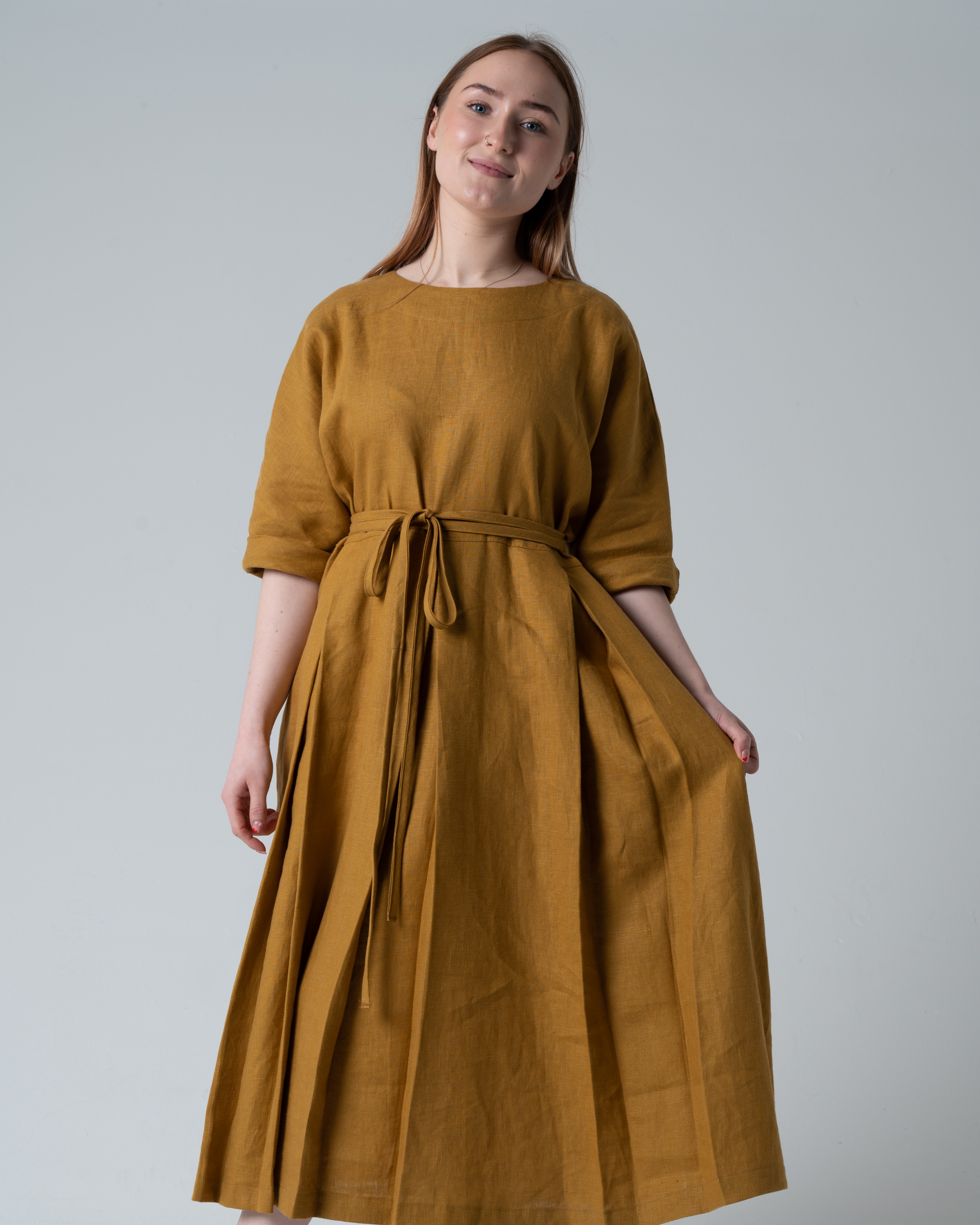 A model wearing golden linen dress with elbow length sleeves and pleated skirt. Standing against a white background.