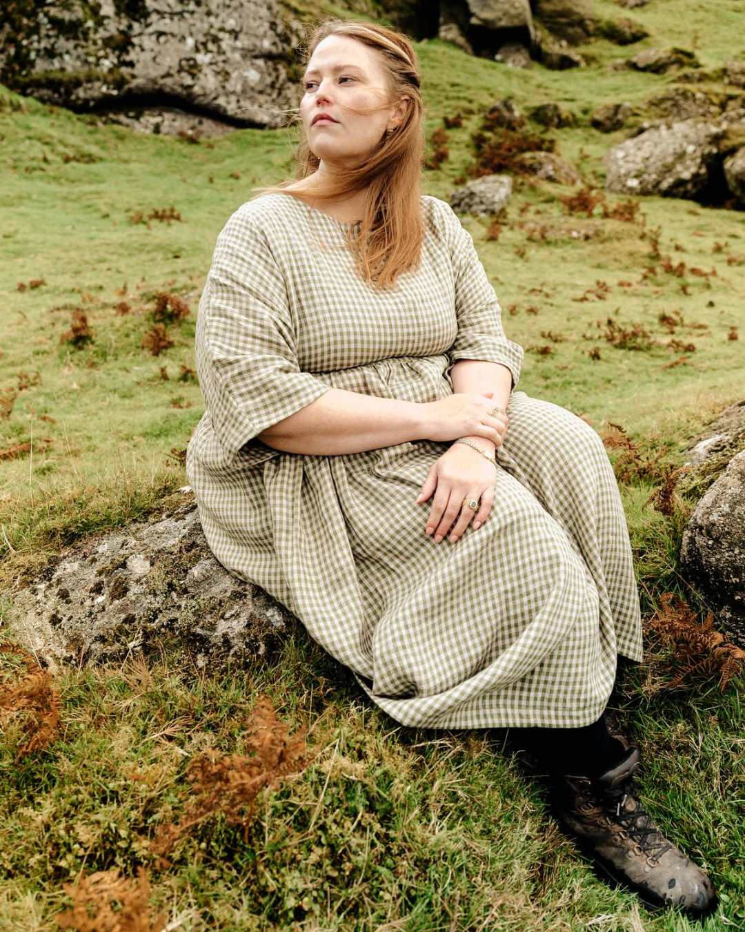 A woman sitting in a natural outdoor setting wearing a green  linen dress with a scooped neckline, short sleeves, and a full skirt. She has a belt with the dress.