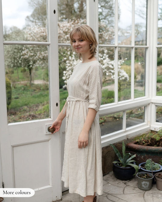 Woman in a beige dress standing in a sunroom with large windows and potted plants.