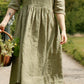 Woman in a green checkered dress standing on a path with a basket, surrounded by nature.